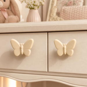 May include: Two cream-colored butterfly-shaped drawer knobs attached to a light-colored wooden dresser. The knobs feature a symmetrical design with detailed wings and a central body, providing a functional handle for opening the drawer fronts.