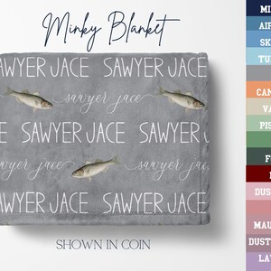 May include: A grey minky blanket with the words "Sawyer Jace" in white, repeated across the surface. The blanket features fish illustrations and the text "Minky Blanket". The colour is shown in "Coin".