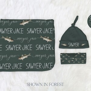 May include: A dark green baby set including a blanket, hat, and headband. The blanket and hat are patterned with the name "Sawyer Jace" in white script and fish. The headband has a fish pattern. The hat says "SAWYER".