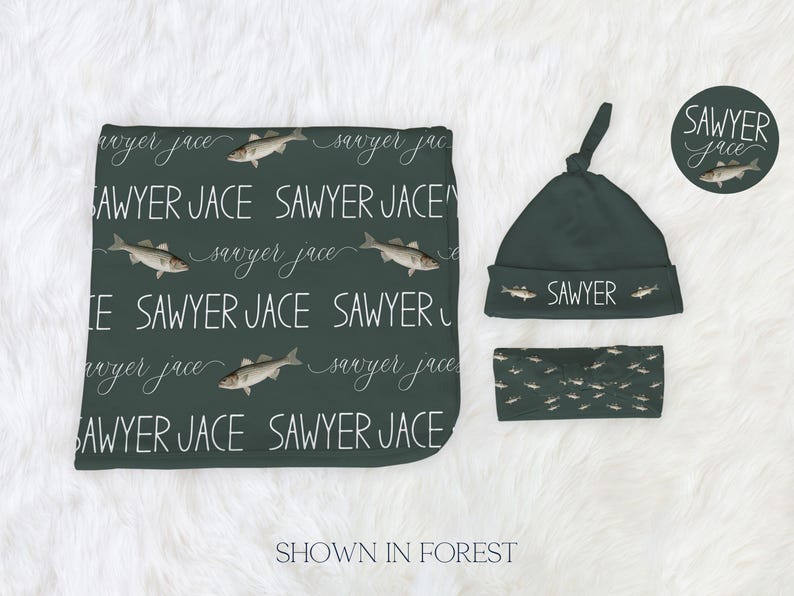 May include: A dark green baby set including a blanket, hat, and headband. The blanket and hat are patterned with the name "Sawyer Jace" in white script and fish. The headband has a fish pattern. The hat says "SAWYER".
