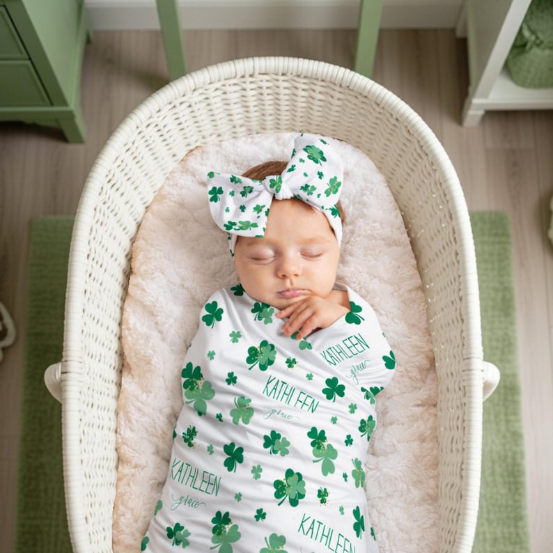 May include: A baby wrapped in a white swaddle blanket with green shamrocks and the name "Kathleen" printed on it. The baby wears a matching headband with a large bow. The baby is in a white wicker bassinet.