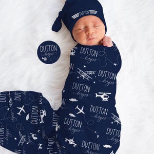 May include: A newborn baby wrapped in a navy blue swaddle blanket and matching cap. The blanket and cap are patterned with white airplanes, helicopters, clouds, and the name "DUTTON hayes". A circular sticker with the same name and airplane graphic is also visible.