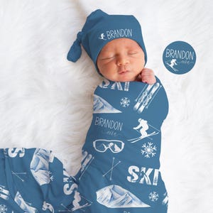 May include: A newborn baby wrapped in a blue swaddle blanket and matching hat. The blanket and hat feature a ski-themed design with mountains, skis, snowflakes, and the name "Brandon" in white. A circular logo with the name "Brandon" and a skier is also visible.