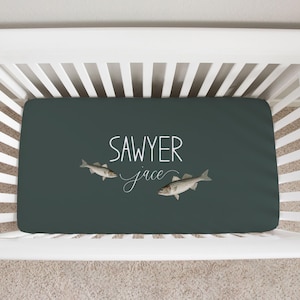 Puede incluir: Una sábana bajera ajustable de color verde oscuro con el nombre "Sawyer" en escritura blanca y dos ilustraciones de peces. El nombre "Jace" está escrito en cursiva debajo de "Sawyer". La sábana se muestra en una cuna blanca.
