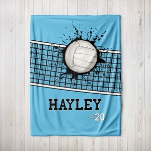 Puede incluir: Una manta de voleibol azul claro con un voleibol blanco y un diseño de salpicaduras negras. La manta presenta un gráfico de red de voleibol y el nombre "HAYLEY" con el número "20".