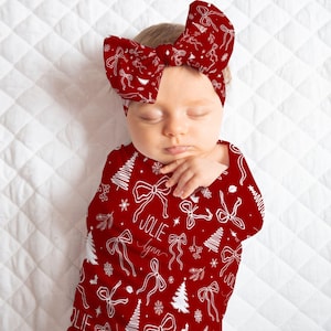 May include: A sleeping baby wrapped in a red swaddle blanket and matching bow headband. The blanket and headband are decorated with white bows, snowflakes, and Christmas trees. The name "Jolie Lynn" is written on the blanket.
