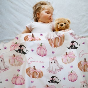 May include: A baby sleeping under a white blanket with a repeating pattern of pink pumpkins, witch hats, ghosts, and candy corn. The name "Gianna" is written on the blanket in pink script. A teddy bear is next to the baby.
