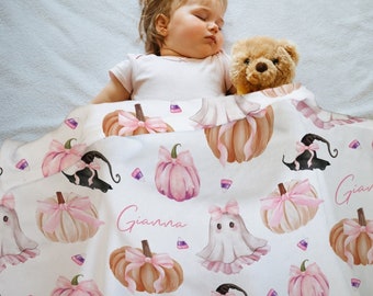 May include: A baby sleeping under a white blanket with a repeating pattern of pink pumpkins, witch hats, ghosts, and candy corn. The name "Gianna" is written on the blanket in pink script. A teddy bear is next to the baby.