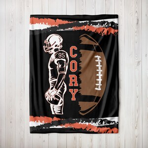 Puede incluir: Manta de franela negra y marrón con una silueta blanca de un jugador de fútbol americano y un balón de fútbol. La camiseta del jugador tiene el número 95. El nombre "Cory" está escrito verticalmente en la manta.