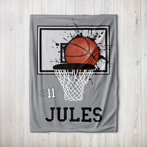 Puede incluir: Una manta gris con temática de baloncesto que muestra un balón de baloncesto pasando por una red. El diseño incluye un aro de baloncesto con un marco blanco y negro, salpicaduras de pintura negra, el número 11 y el nombre JULES en letras negras.