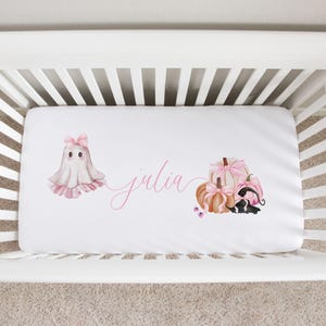 May include: White baby crib sheet with a watercolor design featuring a ghost with a pink bow, pumpkins with pink bows, and the name "julia" in pink script. The sheet is in a white crib.