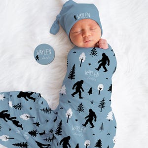 May include: A baby swaddled in a blue blanket and wearing a matching hat. The blanket and hat feature a repeating pattern of black Bigfoot silhouettes, trees, and the text "WAYLEN banks". A round sticker with the same design is also present.