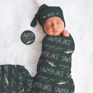May include: A newborn baby swaddled in a dark green blanket and matching knotted hat. The blanket and hat feature the name "Sawyer Jace" in white script and fish illustrations. A round sticker with the same design is also visible.