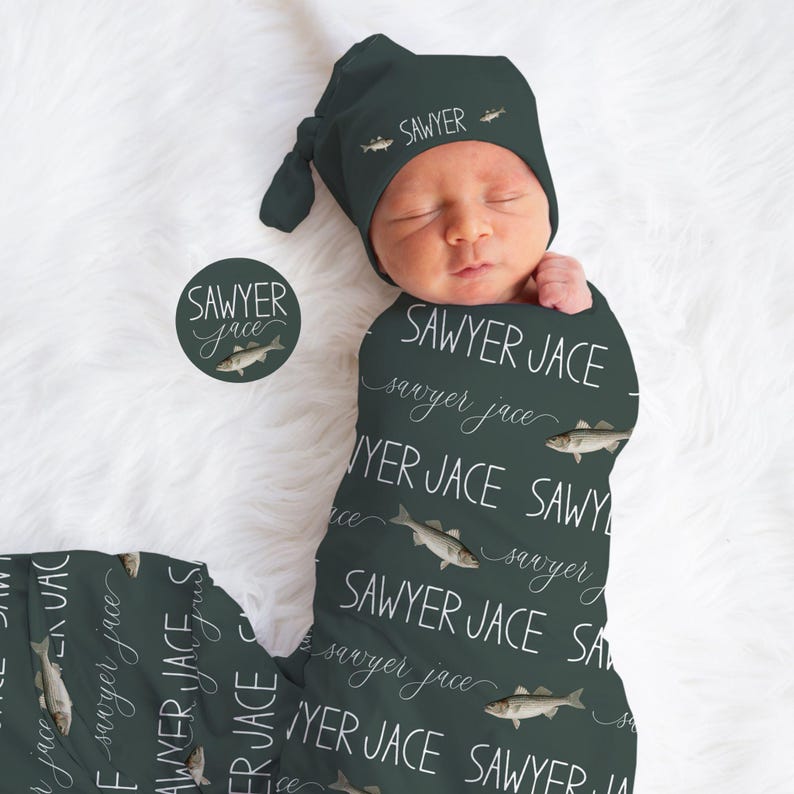 May include: A newborn baby swaddled in a dark green blanket and matching knotted hat. The blanket and hat feature the name "Sawyer Jace" in white script and fish illustrations. A round sticker with the same design is also visible.