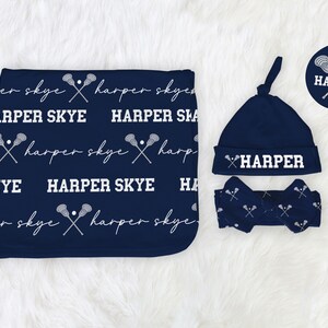 May include: A navy blue baby blanket, hat, and headband set. The blanket features the text "HARPER SKYE" and lacrosse stick graphics. The hat and headband also have the name "HARPER" in white. The set is on a white, fluffy surface.