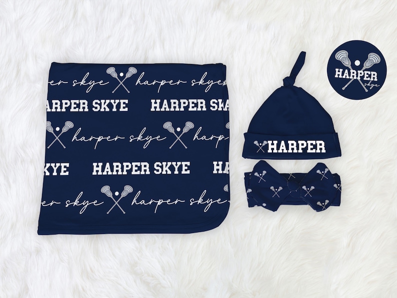May include: A navy blue baby blanket, hat, and headband set. The blanket features the text "HARPER SKYE" and lacrosse stick graphics. The hat and headband also have the name "HARPER" in white. The set is on a white, fluffy surface.