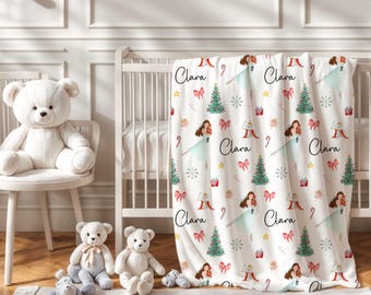 May include: A white fleece blanket featuring a repeating Christmas pattern of green pine trees, red gift boxes, pink bows, candy canes, yellow stars, and illustrations of a girl in a light blue dress, with the name Clara printed in black script.