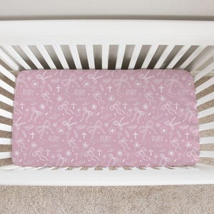 May include: A fitted crib sheet with a pink background and a repeating pattern of white bows, crosses, flowers, and the name "RUBY claire". The sheet is designed to fit a standard crib.