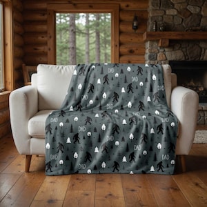 May include: A grey blanket featuring a pattern of black Bigfoot figures, trees, and the word "DAD" in white. The blanket is draped over a white sofa in a rustic living room setting.