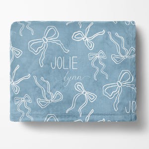 May include: A soft, blue blanket with a repeating pattern of white bows. The name "JOLIE Lynn" is printed in white script. The blanket is made of a plush material.