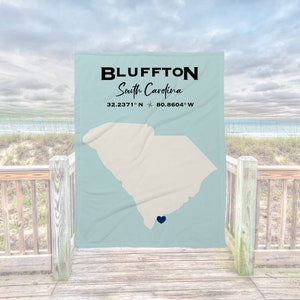 May include: A light blue throw blanket with a white outline of the state of South Carolina and a blue heart marking the location of Bluffton, South Carolina. The text "Bluffton, South Carolina 32.2371° N 80.8604° W" is printed on the blanket.