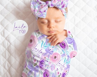 May include: A swaddle blanket and matching headband set featuring a white background with a repeating pattern of purple and pink watercolor roses, light purple bows, and teal leaves. The text Laila Rae is printed throughout the fabric.