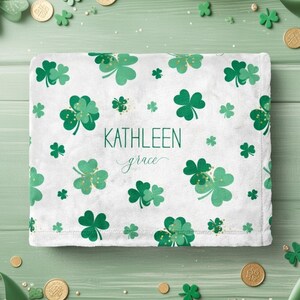 May include: White blanket with a pattern of green shamrocks and gold accents. The name "KATHLEEN grace" is printed in green. The blanket is surrounded by gold coins and shamrock decorations, creating a festive St. Patrick's Day theme.