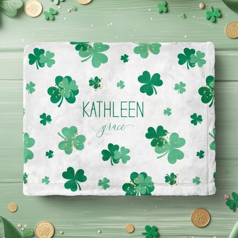May include: White blanket with a pattern of green shamrocks and gold accents. The name "KATHLEEN grace" is printed in green. The blanket is surrounded by gold coins and shamrock decorations, creating a festive St. Patrick's Day theme.