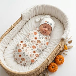 May include: A newborn baby swaddled in a white fabric wrap featuring a repeating pattern of orange pumpkins, brown flowers, and the name Willow Sage in black script, paired with a matching white knotted hat labeled with a small pumpkin and Willow.