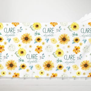Puede incluir: Manta de bebé blanca con un estampado repetido de girasoles amarillos, flores naranjas y hojas verdes. El nombre "CLARE Elizabeth" está escrito en escritura verde oscuro. La manta está sobre una cuna blanca.