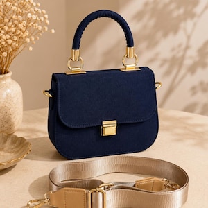 May include: A navy blue handbag with a rounded base and a flap closure secured by a gold-toned metal clasp. The bag features a matching navy blue top handle with gold hardware and a detachable, adjustable beige woven shoulder strap with tan accents.