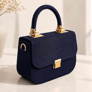 May include: A structured navy blue handbag with a matte finish, featuring a single top handle, gold-tone metal hardware, a rectangular flap closure with a gold clasp, and side D-rings for an optional shoulder strap attachment.