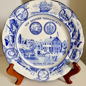 May include: A white ceramic plate with a blue transferware design featuring a scene of New Bedford, Massachusetts. The plate displays a ship, two circular seals, and the text New Bedford Massachusetts and New Bedford Fifty Years Ago.