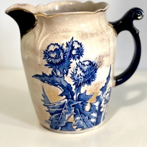 May include: Cream-colored ceramic pitcher with a blue thistle floral design. The pitcher has a black handle and spout, with gold trim around the rim. The floral design features blue thistles and leaves.