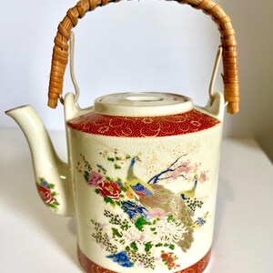 May include: Cream-colored ceramic teapot with a woven handle. The teapot features a red patterned band at the top and bottom, and a detailed illustration of peacocks and flowers. The handle is made of woven brown material.