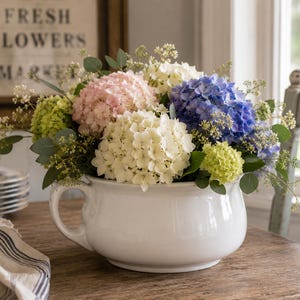 May include: A white ceramic flower pot filled with a vibrant arrangement of hydrangeas in shades of pink, blue, white, and green. The pot has a handle and sits on a wooden table. A sign that says "FRESH FLOWERS MARKET" is in the background.