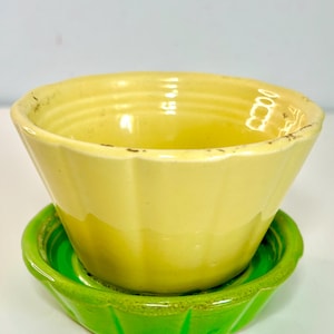 May include: A yellow ceramic bowl with a fluted design sits atop a bright green saucer. The bowl has a slightly distressed appearance, with visible wear on the rim. The set is likely vintage or vintage-inspired, suitable for serving or display.