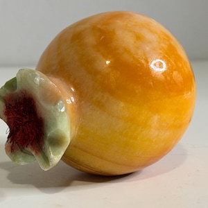 May include: A polished, rounded stone sculpture shaped like a pomegranate. The body is a translucent, marbled golden-orange hue. The top features a pale green, carved calyx with a textured, dark red fibrous center, mimicking the fruit's crown.