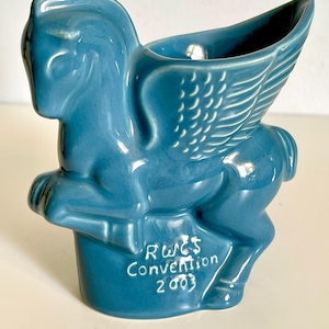 Puede incluir: Figura de Pegaso de cerámica azul con alas detalladas. La figura tiene una pequeña abertura en la parte posterior y el texto "RWE'S Convention 2003" está impreso en la base. El Pegaso es una criatura mítica.