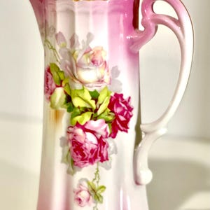 Puede incluir: Una cafetera alta de porcelana vintage con tapa, con un diseño degradado rosa y blanco. La cafetera está adornada con un motivo floral de rosas en tonos rosa, rojo y verde, con detalles dorados.