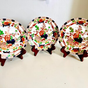 May include: Three round ceramic plates featuring a white base with a colourful floral pattern in red, green, and blue. Each plate has a dark blue and white geometric border along the rim. The plates are displayed upright on small brown stands.