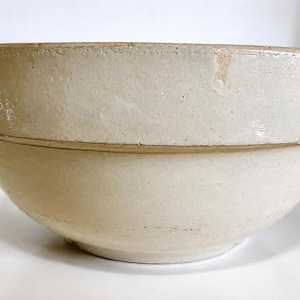 May include: A large, round, off-white ceramic mixing bowl with a thick, prominent rim and a smooth, glazed finish. The bowl features a wide, deep basin and a slightly tapered base, showing subtle variations in the light-coloured surface texture.