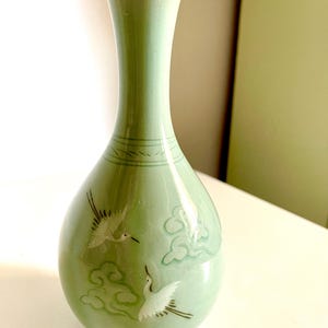 May include: A pale green ceramic vase with a slender neck and rounded body. The vase features a decorative design of white cranes and stylized clouds. The vase is approximately 20 cm tall, making it a decorative accent piece.