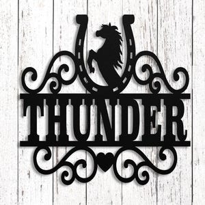 May include: Black metal wall art with a horseshoe and horse silhouette above the word "THUNDER". A horse with a white mane and tan face looks out from a stable window. The background is a white wooden wall.