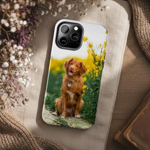 May include: A white phone case featuring a brown dog with a yellow flower field background. The dog is wearing a heart-shaped tag. The case has a black camera module and is resting on a beige surface with flowers and a book.
