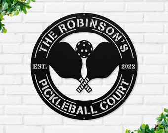 May include: Black metal sign with the text "The Robinson's Pickleball Court" and the years "EST. 2022". Two crossed pickleball paddles and a ball are in the center of the sign.