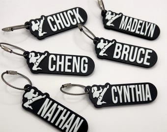 May include: Black luggage tags with white text and a martial arts figure. Each tag displays a different name: CHUCK, MADELYN, BRUCE, CHENG, NATHAN, and CYNTHIA. The tags have a metal loop for attachment.
