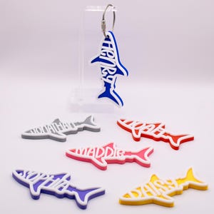 May include: Shark-shaped keychains in various colors, each with a name cut out. The keychains are blue, gray, red, pink, purple, and yellow with white accents. One keychain has a metal loop for hanging.