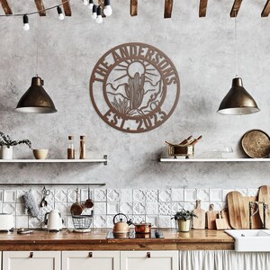 May include: A brown wooden sign with a circular design featuring a desert landscape with a cactus and the text "The Andersons Est. 2023". The sign is hanging on a gray wall in a kitchen setting.