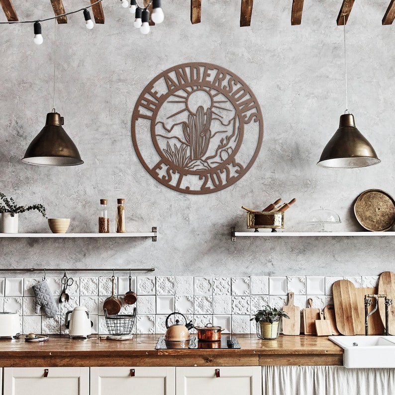 May include: A brown wooden sign with a circular design featuring a desert landscape with a cactus and the text "The Andersons Est. 2023". The sign is hanging on a gray wall in a kitchen setting.